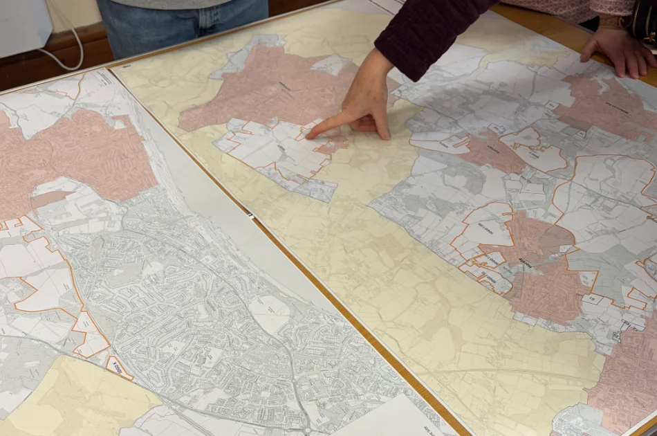 A person points to a location on a large map spread across a table, whilst another person stands nearby. Multiple maps with coloured and outlined regions are visible.